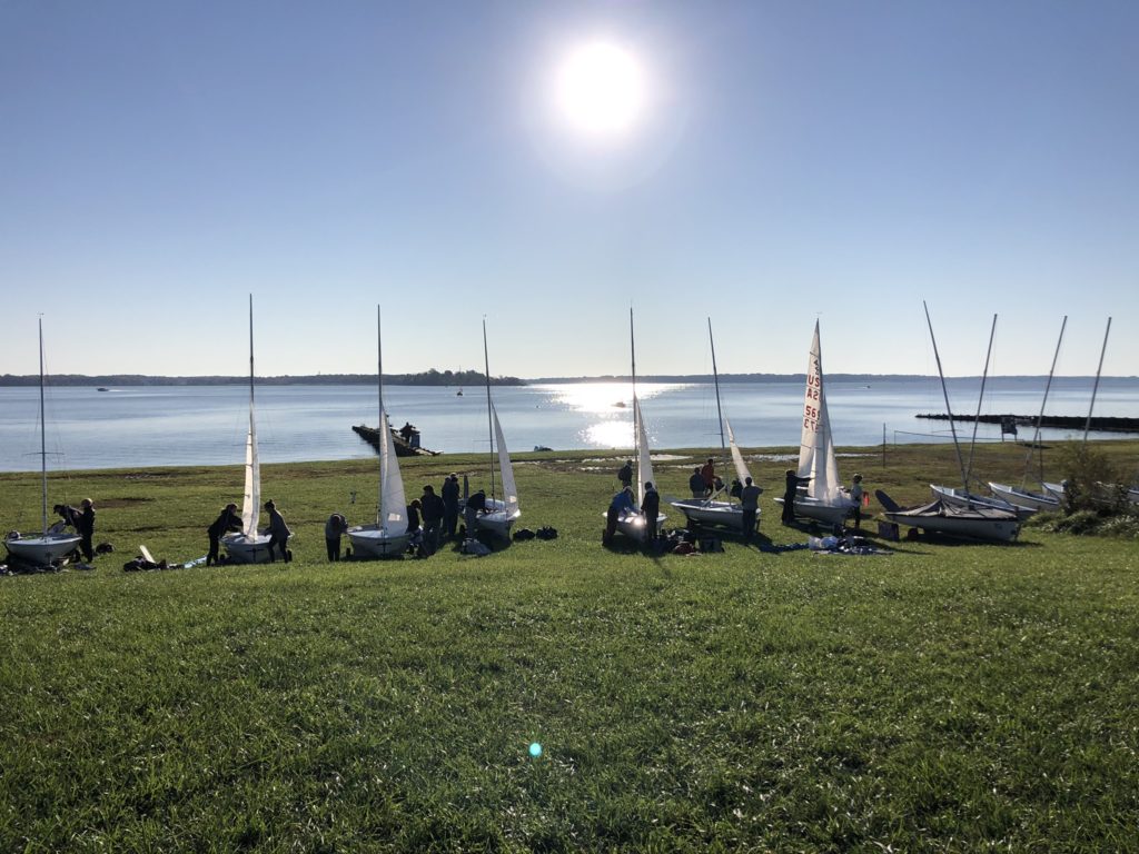 A group of small sailboats with people preparing them on a grassy hill by a calm lake. The sun shines brightly in the clear blue sky, reflecting off the water.