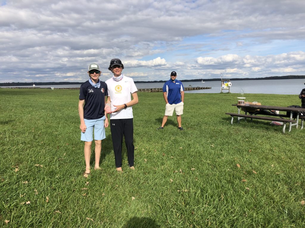 Two people stand on grass holding an award, smiling at the camera, with a third person in a blue shirt standing behind them. A lake, picnic table, and cloudy sky are visible in the background.