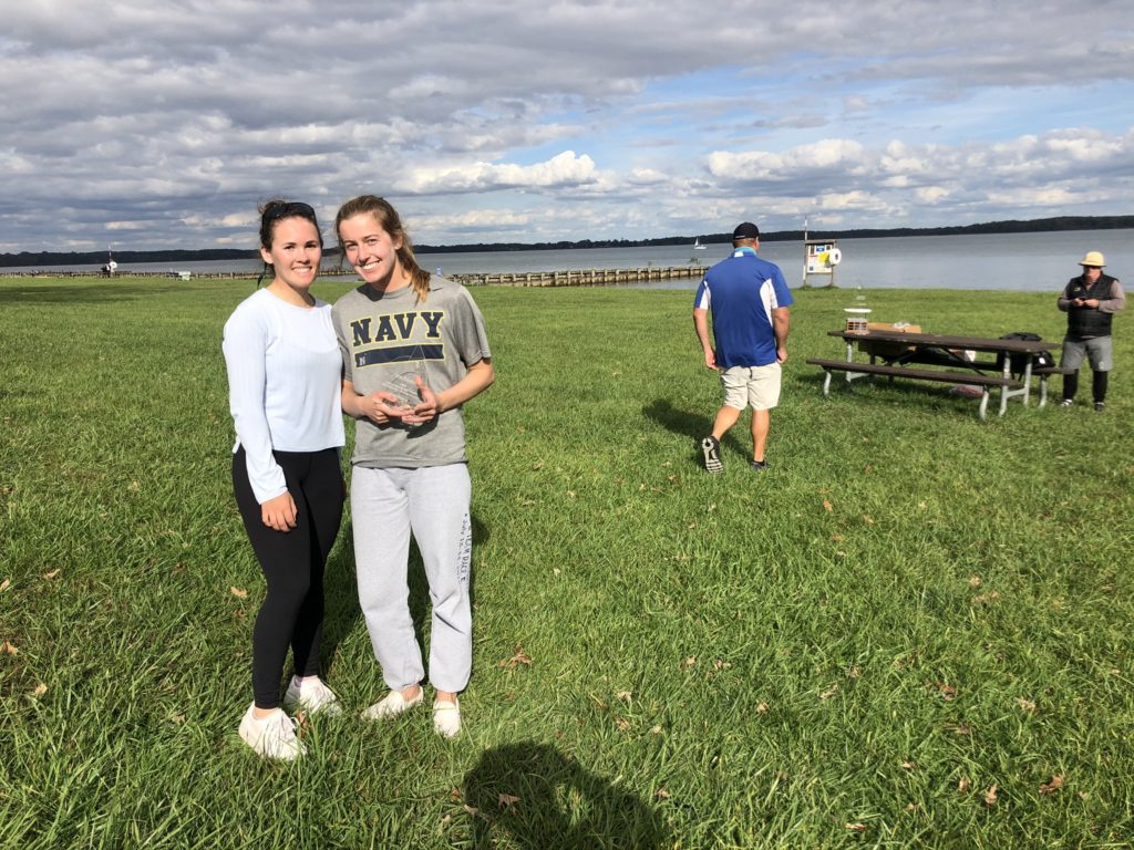 Two young women stand and smile on a grassy field near a lake, one wearing a Navy t-shirt. In the background, people are near a picnic table and dock under a partly cloudy sky.