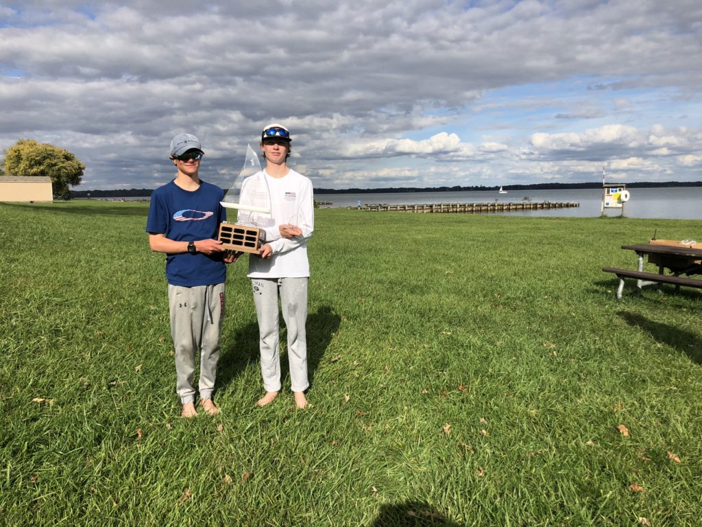 Two people in casual athletic clothes stand on grass by a lake, holding a trophy shaped like a sailboat. A wooden pier, picnic table, and partly cloudy sky are visible in the background.