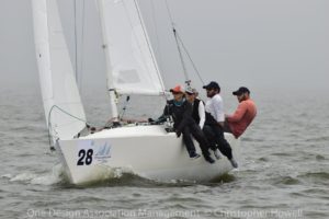 Four people sail a white racing sailboat numbered 28 on calm, gray water. Three sit on the edge leaning out, while the fourth steers. All wear hats and sailing gear. The sky appears overcast.