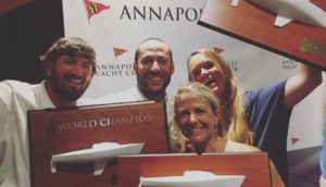 Four smiling people pose together holding trophies shaped like sailboats in front of an Annapolis Yacht Club backdrop. A "World Champion" plaque is visible. They appear to be celebrating a sailing victory.