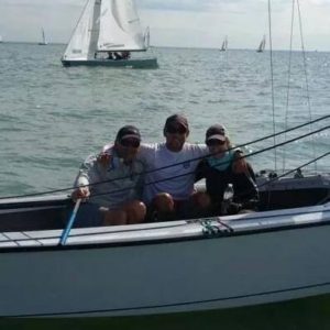 Three people sit together, smiling, on a small sailboat in the water. Several other sailboats are visible in the background under a partly cloudy sky.