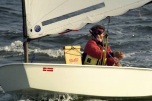 A person wearing a red life jacket and sunglasses sails a small white dinghy with a Danish flag sticker, smiling and holding the tiller on choppy water. A box labeled "STALKER" is secured inside the boat.