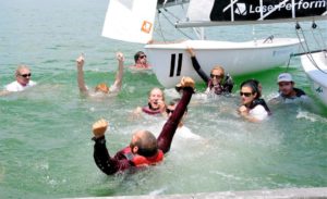 A group of people in life jackets celebrate in the water next to a sailboat on a sunny day, some raising their arms and smiling joyfully.