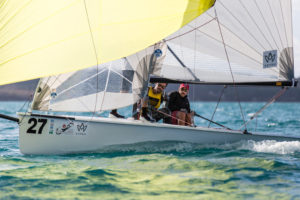 A small sailboat with the number 27 glides across blue water. Two people, wearing hats and sunglasses, steer the boat as its white and yellow sails catch the wind. The background is blurred, focusing on the sailing action.