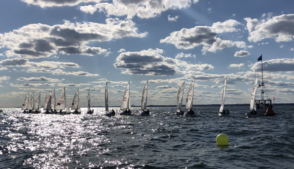 A group of small sailboats with white sails line up on a sparkling, sunlit body of water under a sky filled with scattered clouds. A buoy and a boat with a flag are visible on the right.