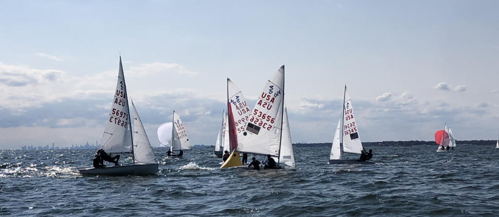 Several small sailboats with white sails, marked with numbers and "USA," race on choppy water under a partly cloudy sky. The distant city skyline and more boats are visible on the horizon.
