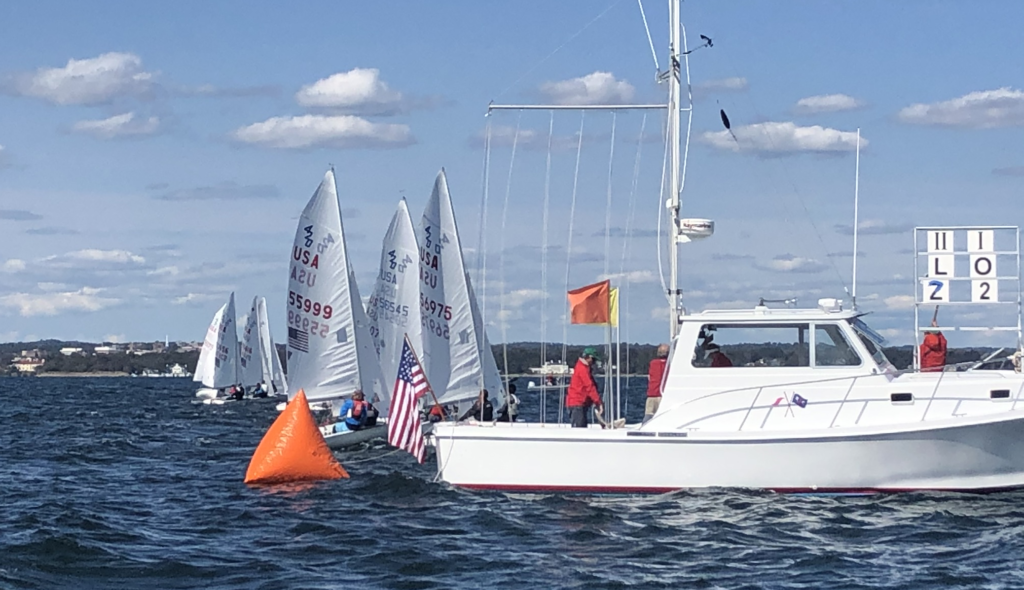 Several sailboats line up near an orange buoy and a white boat with race officials, flags, and scoreboards on a sunny day with blue water, gentle waves, and a shoreline in the distance.