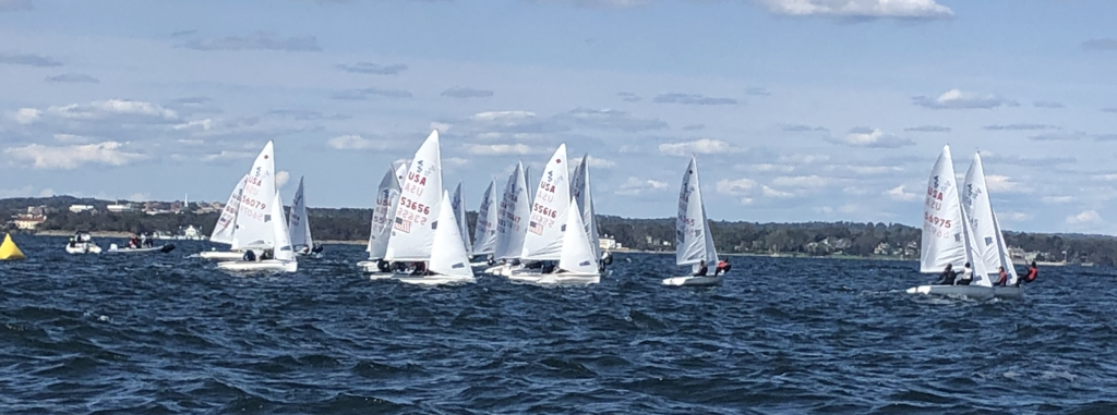 A group of small sailboats with white sails race on choppy blue water under a partly cloudy sky. Distant shorelines and trees are visible in the background. A yellow buoy and a motorboat can be seen to the left.
