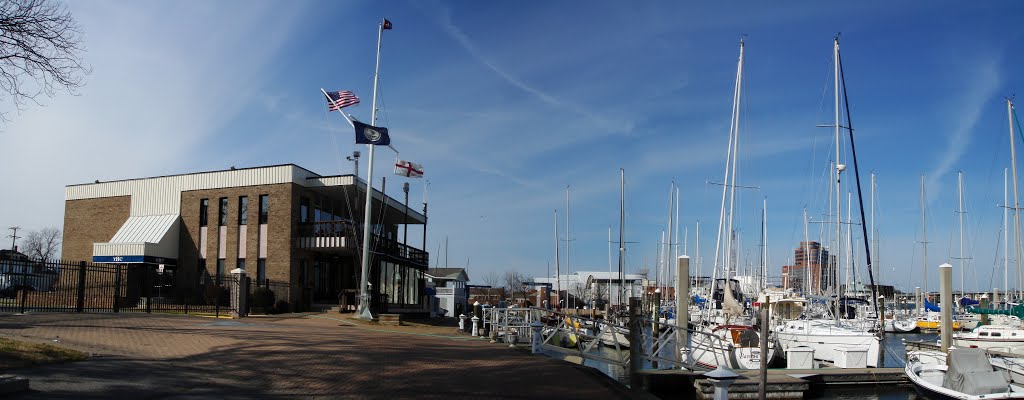 A marina with sailboats docked near a brown brick building flying several flags, under a clear blue sky. A city skyline is visible in the background. The scene is calm and sunny.
