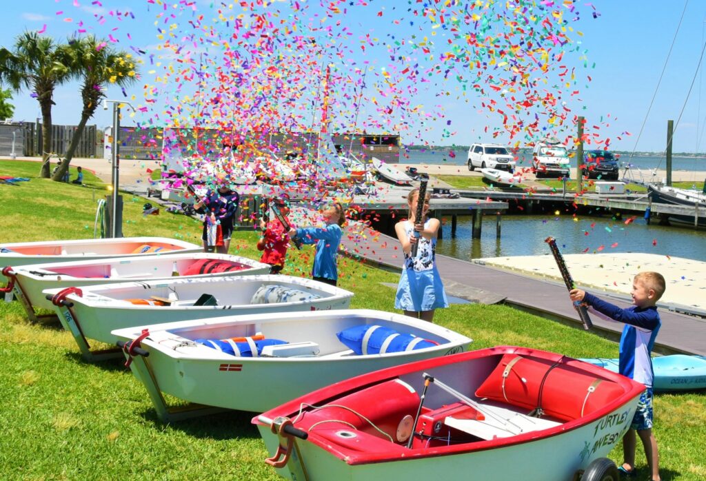 Children celebrate by launching colorful confetti into the air beside small sailboats on a grassy area near a marina, with water, docks, and palm trees in the background under a sunny sky.