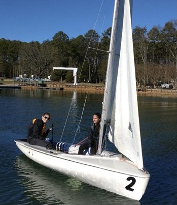 Two people sit in a small white sailboat with the number 2 on the bow, floating on calm water near a wooded shore under a clear blue sky.
