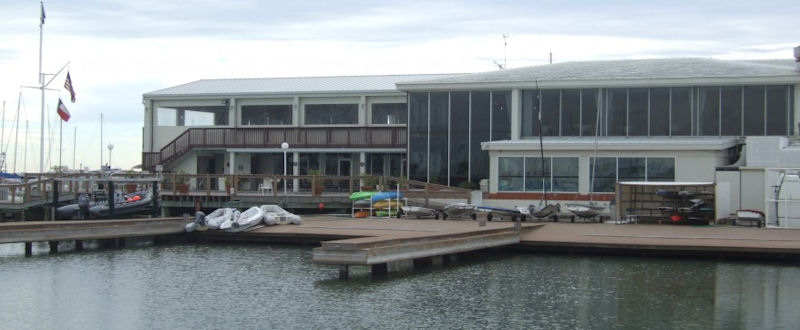 A waterfront building with large windows, wooden decks, and boats docked alongside. Several flags are flying on the left, and the water is calm under a cloudy sky.