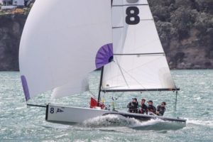 A sailboat with the number 8 on its sail glides swiftly on the water with four people onboard, wearing life jackets and sailing gear. The boat is near a rocky, tree-covered shoreline.