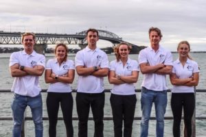 Six young adults stand side by side in matching white polo shirts and black pants, arms crossed, in front of a body of water with a large bridge in the background on a cloudy day.