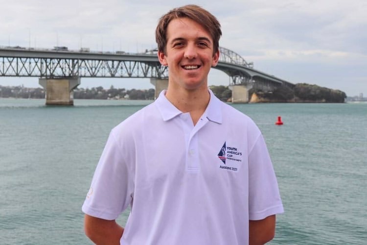 A young man in a white polo shirt stands smiling in front of a body of water, with a large bridge and cloudy sky in the background.