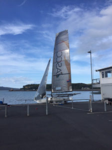 A small sailboat with tall sails is parked on a waterfront dock under a partly cloudy blue sky, with calm water and green hills in the background.
