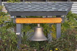 A large metal bell hangs beneath a wooden sign that reads "Mission Bay Yacht Club," surrounded by green plants and flowers. The structure has a shingled roof.