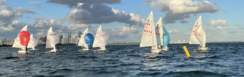 Small sailboats with colorful sails race on choppy water under a partly cloudy sky, with a city skyline and tall buildings visible in the background. A yellow buoy floats nearby.