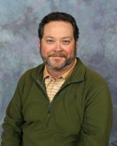 A man with short dark hair and a beard, wearing a green zip-up jacket over a beige striped shirt, sits and smiles in front of a blue and gray mottled background.
