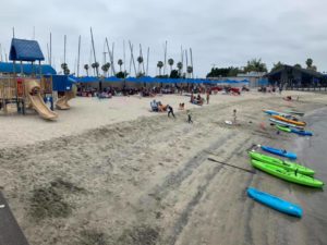 A sandy beach scene with people relaxing under blue canopies, children playing near a playground, and colorful kayaks lined up at the water’s edge. Sailboats and palm trees are visible in the background under a cloudy sky.
