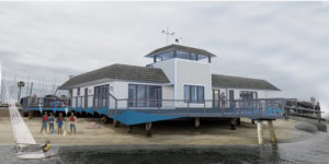A modern boathouse with a gray roof and blue railings stands on stilts above the sand. People gather on the shore and deck, while a small sailboat is in the water under a cloudy sky.