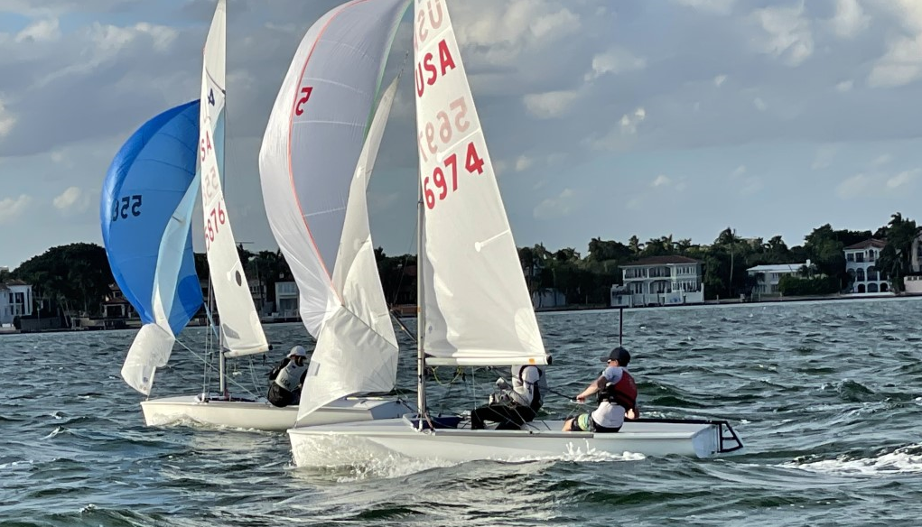 Two small sailboats with white sails and 'USA' markings race side by side on a choppy body of water, with people actively maneuvering the boats. Houses and trees line the distant shore under a partly cloudy sky.