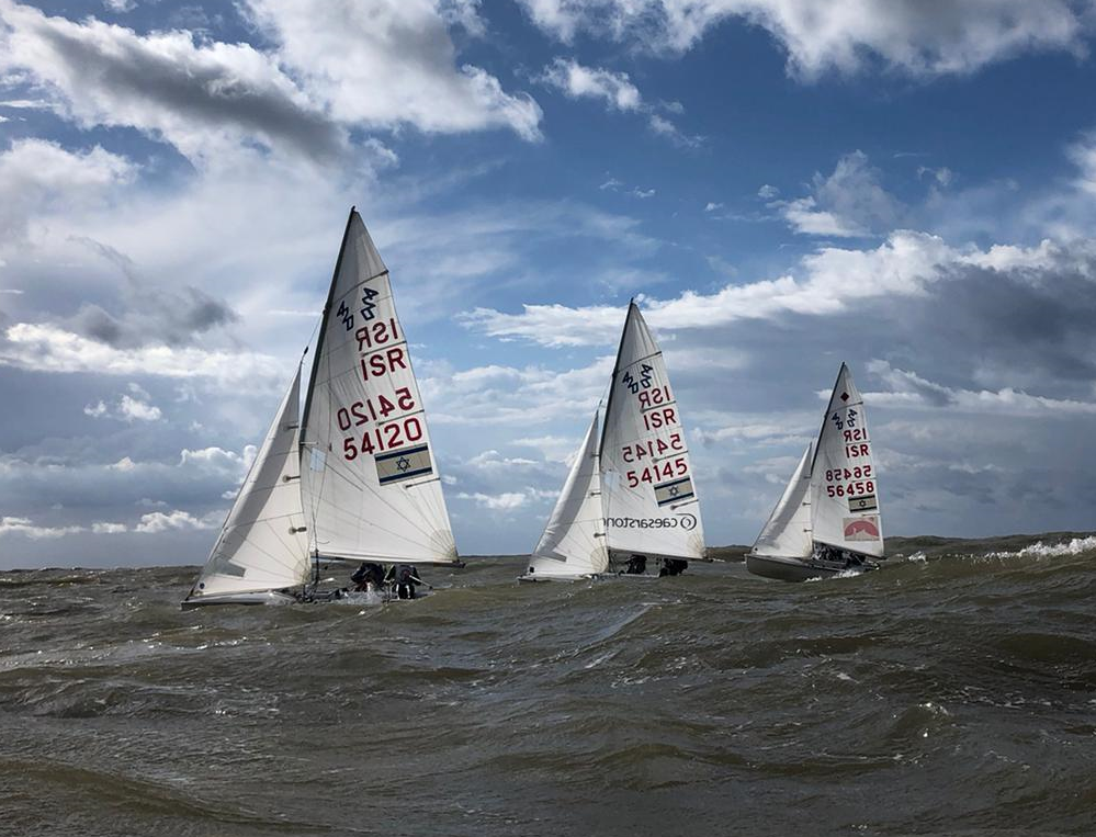 Three small sailboats with white sails and red numbers race on choppy water under a partly cloudy sky. The sails display various codes, and the boats are closely grouped, suggesting an active sailing competition.