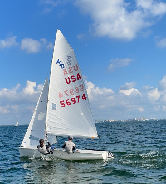 Two people sail a small white sailboat with USA markings on blue water under a partly cloudy sky. Buildings are visible in the distant background, and another sailboat is seen far away.