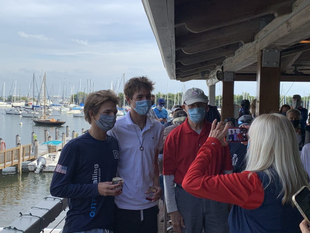 Two young men wearing masks pose for a photo with an older man in red near a marina, while a woman takes their picture. Several boats and docked sailboats are visible in the background.