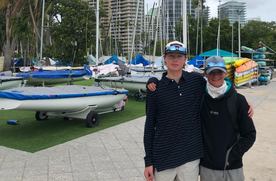 Two people stand arm in arm, smiling at the camera in front of several small sailboats and kayaks stacked by a marina. Tall buildings and trees are visible in the background.