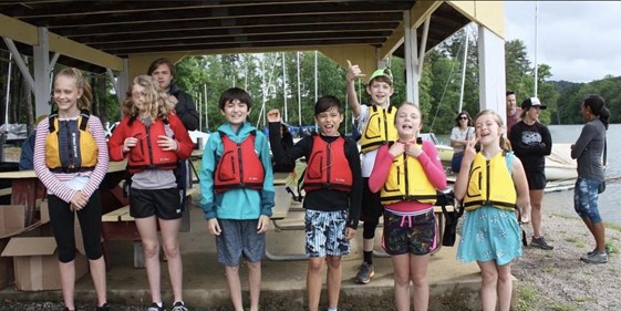 A group of smiling kids in colorful life jackets stand under a shelter by a lake, posing for a photo. Some are making excited gestures, while adults and sailboats are visible in the background.