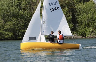 Two people wearing life jackets sail a small yellow sailboat with white sails on a calm body of water, with green trees and foliage visible in the background.