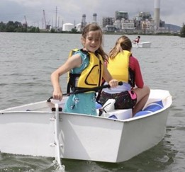 Two girls wearing life jackets are rowing a small white boat on a body of water, with an industrial plant and cranes visible in the background. One girl faces the camera and smiles while rowing.