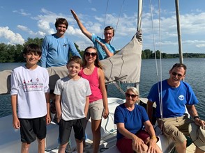 A group of seven people, including adults and children, smile and pose on a sailboat with a lake and trees in the background on a sunny day.