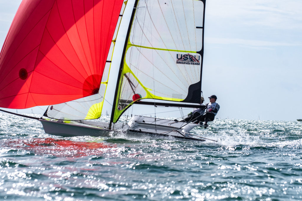 A person steers a small sailboat with bright red and yellow sails on open water, creating splashes as they race. The sail displays "USA Sailing Team." The sky is partly cloudy and the sea is slightly choppy.