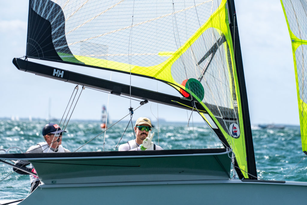 Two sailors wearing hats and sunglasses navigate a sailboat with neon yellow sails on choppy blue water, with other boats and a distant horizon in the background under a partly cloudy sky.