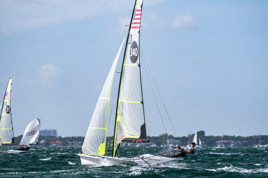 Two sailors lean out from a racing sailboat with the number 340, navigating choppy waters. Another sailboat is seen behind them, with a coastal shoreline and some buildings in the background under a partly cloudy sky.