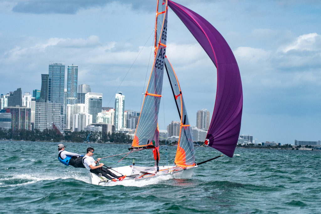 Two people sail a small boat with a large purple sail on choppy water, with a city skyline and tall buildings visible in the background under a partly cloudy sky.