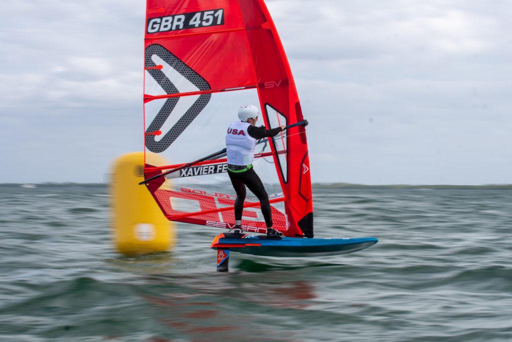 A windsurfer wearing a white helmet and "USA" vest glides on a blue and red sailboard past a yellow buoy on choppy water, under a cloudy sky.
