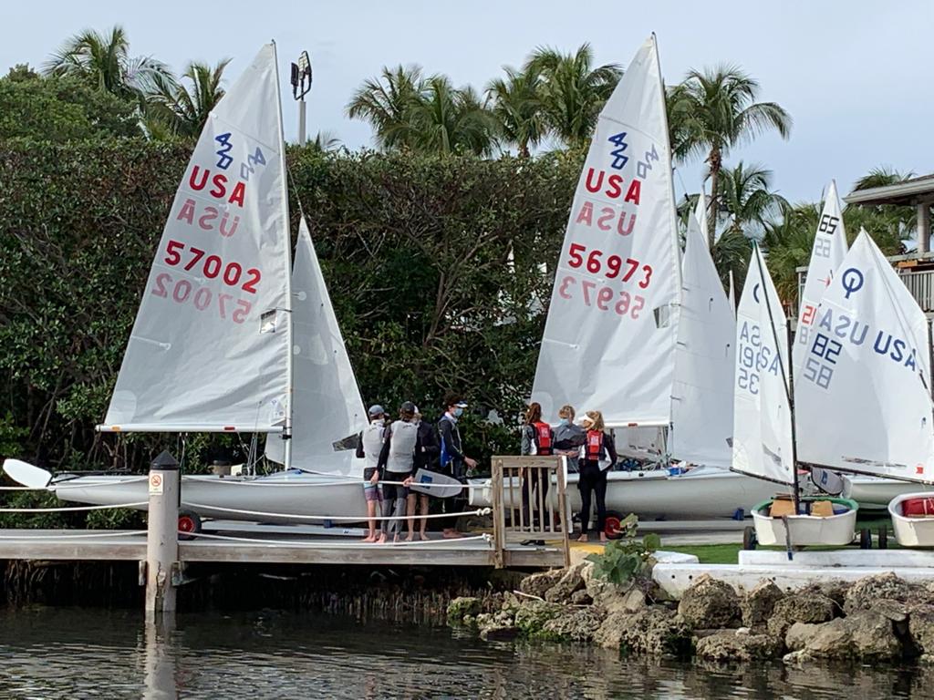 Several people stand on a dock and near the shore next to three sailboats with large sails marked 'USA' and numbers, surrounded by greenery and palm trees near the water.