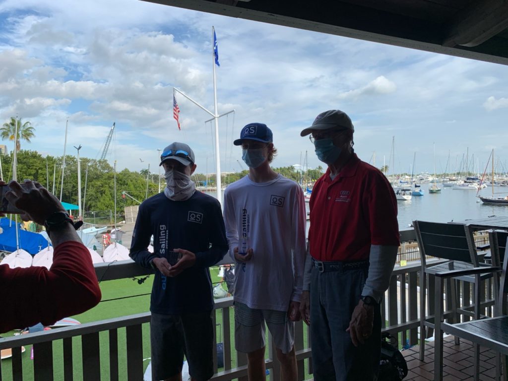 Three people stand together on a balcony overlooking a marina with sailboats. Two young people in sailing gear and masks hold trophies, while an older man in a red shirt and cap stands beside them.