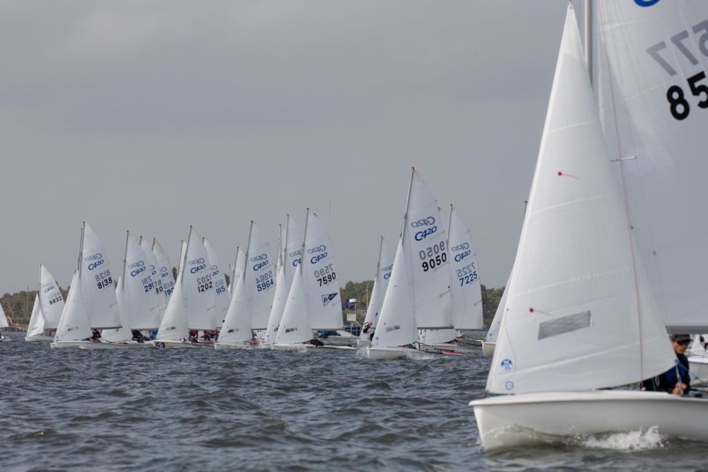 A group of small sailboats with white sails line up on a choppy body of water, racing closely together under a cloudy sky. Sail numbers and logos are visible on the sails.
