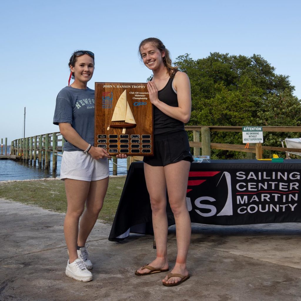 Two young women stand outdoors by the water, smiling and holding a large wooden sailing trophy. A table with a “Sailing Center Martin County” banner is beside them, and trees and a dock are in the background.