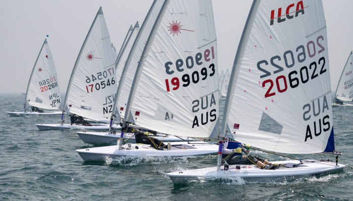A group of sailors competes in a dinghy race on the ocean, each maneuvering small white sailboats with numbers and country codes on the sails. The water is slightly choppy and the sky is hazy.