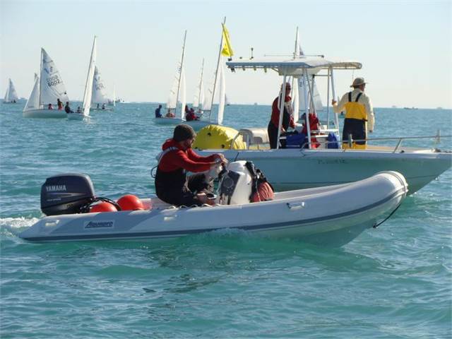 A person drives an inflatable motorboat on calm water, passing a white motorboat with people standing on it. Several sailboats and a yellow buoy are visible in the background.