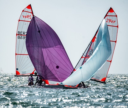 Two small sailboats with colorful sails—one purple, one light blue—are close together on choppy water, with sailors visible aboard each boat under a bright sky.