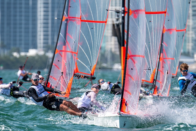 Sailors compete in a high-speed dinghy race, leaning out to balance their boats as water splashes. Tall buildings are visible in the background under a clear sky.