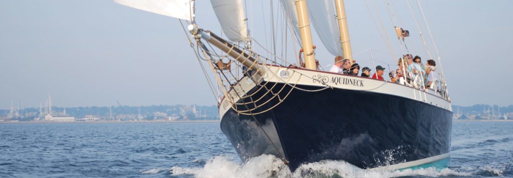 A large sailboat named “Aquidneck” sails on calm water with people onboard enjoying the ride; the shoreline is visible in the background under a hazy sky.